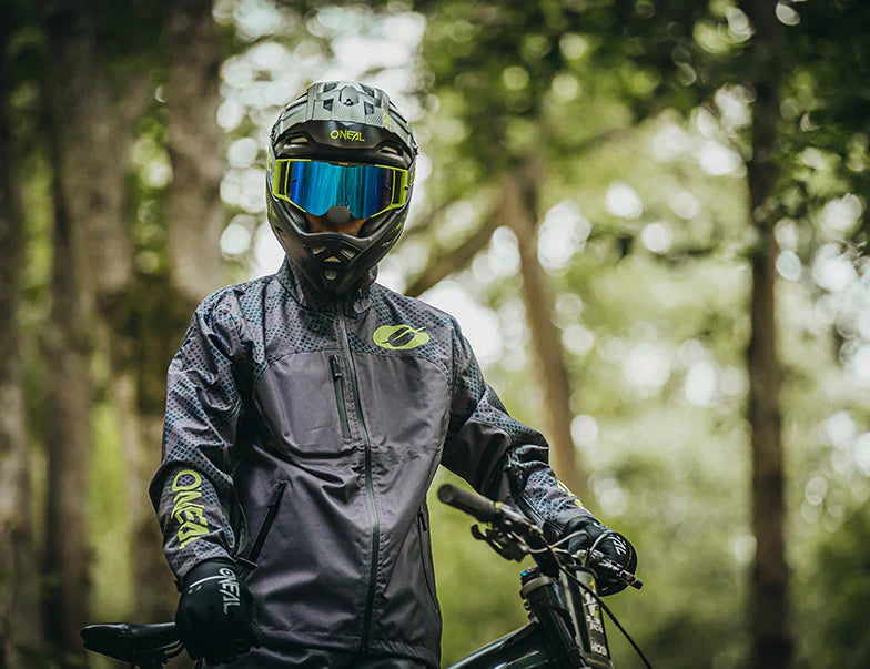Mężczyzna w odzieży przeciwdeszczowej O'NEAL na rowerze górskim w lesie, gotowy na jazdę w deszczu.